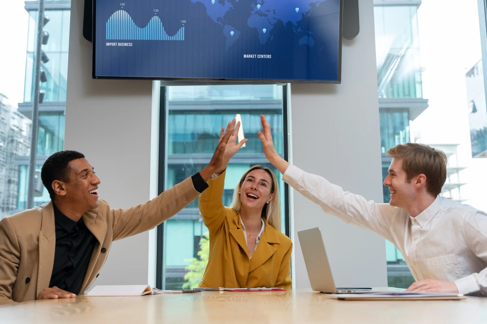 Três colegas de trabalho sorridentes sentados em uma mesa de reunião celebram com um "high five" coletivo. Ao fundo, há janelas de vidro com vista para um prédio moderno e uma tela com gráficos e mapa mundial, indicando dados de negócios. A cena transmite colaboração, entusiasmo e sucesso em um ambiente corporativo moderno.