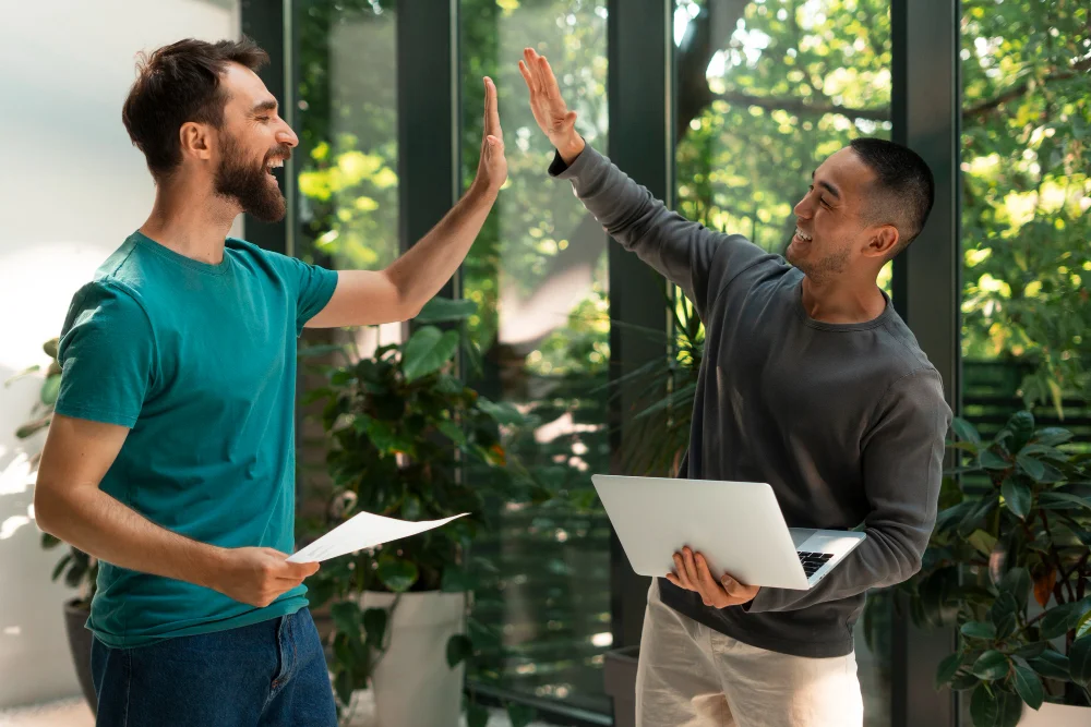 Dois homens em um ambiente iluminado e cercado por plantas trocam um "high five" com expressões de alegria. Um deles segura uma folha de papel, enquanto o outro carrega um notebook. Ambos estão sorrindo, em clima descontraído, sugerindo comemoração ou sucesso em uma tarefa profissional.
