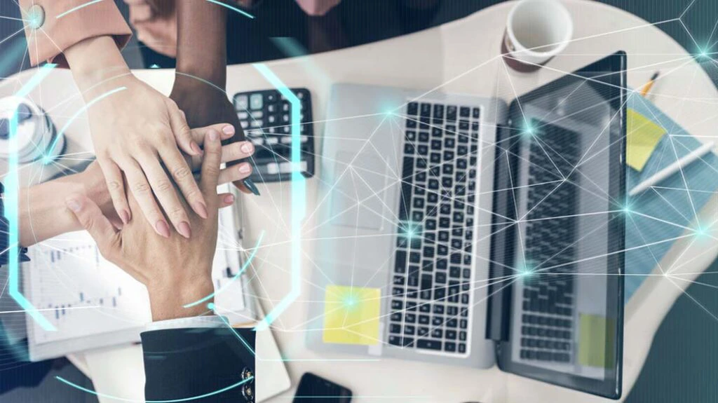 Vista superior de uma mesa de trabalho com notebook, calculadora, gráficos e caneca, onde várias mãos de pessoas de diferentes tons de pele estão sobrepostas em sinal de união e colaboração. A imagem possui elementos gráficos de conexões digitais e tecnologia, representando inovação e trabalho em equipe.