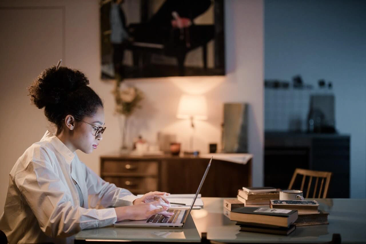 Mulher jovem com óculos e cabelo preso trabalha concentrada em um laptop, sentada à mesa em um ambiente doméstico aconchegante e levemente iluminado por um abajur ao fundo. Há livros, um caderno e uma xícara sobre a mesa, sugerindo um momento de estudo ou autoaprendizagem.