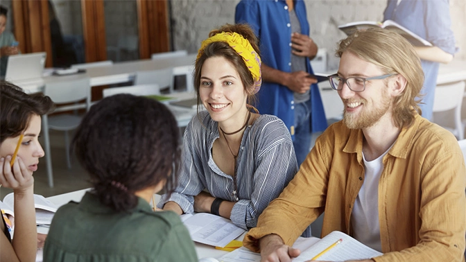 amigos em processo de aprendizagem felizes todos sentados em uma mesa, apenas dois de frente um homem de amarelo e loiro e uma melhor feliz de cinza e faixa amarela na cabeça