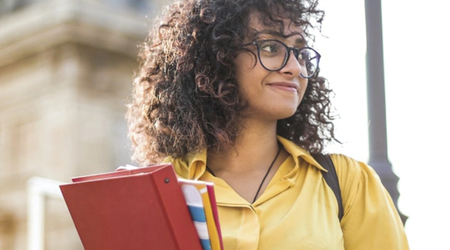 Mulher jovem de cabelos cacheados e óculos sorri levemente enquanto caminha ao ar livre durante o dia. Ela veste uma camisa amarela e carrega alguns livros e cadernos coloridos nos braços. Ao fundo, há uma construção histórica desfocada e um poste de iluminação.