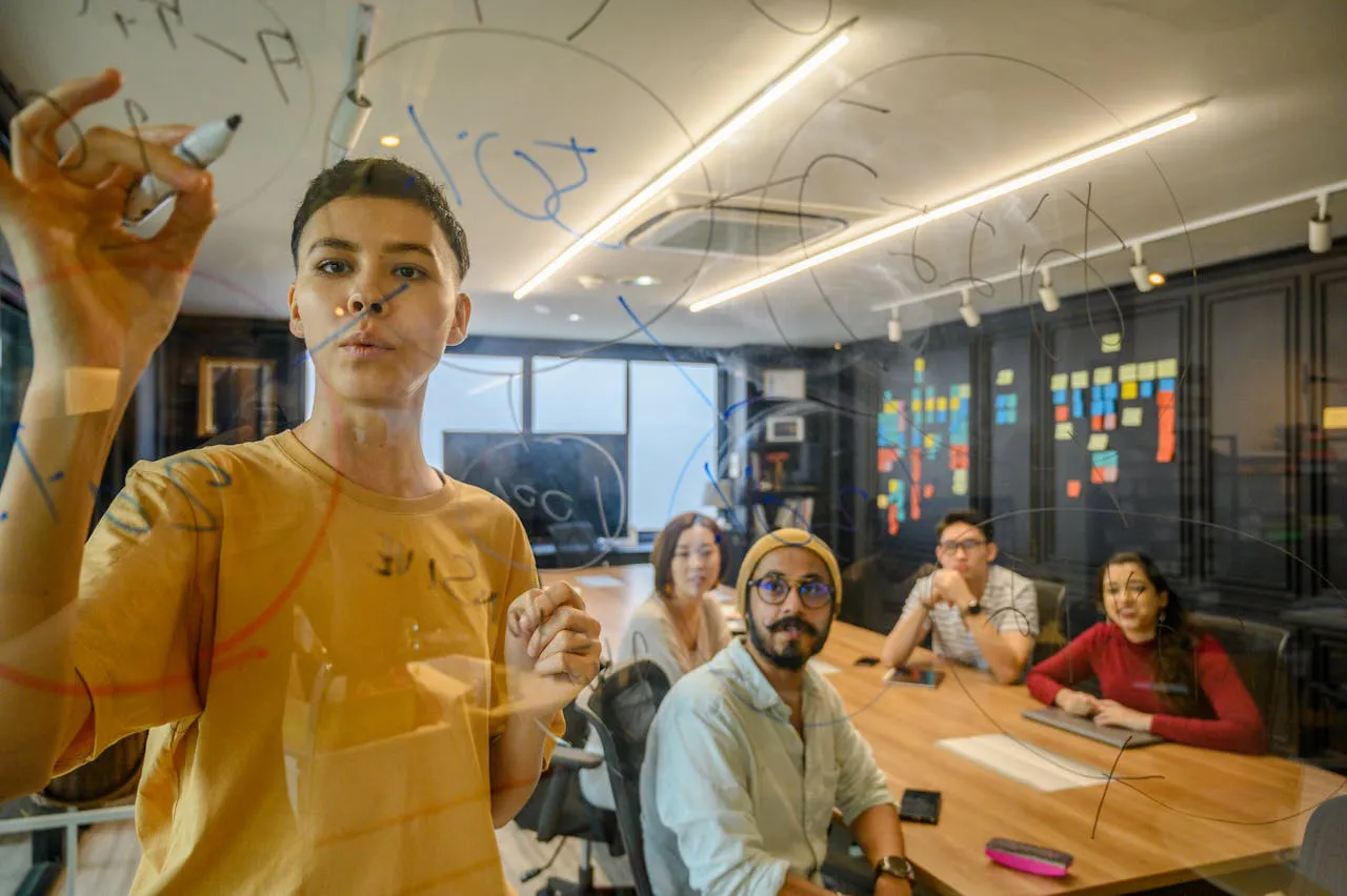 Um jovem com camiseta amarela escreve em um painel de vidro transparente com um marcador enquanto apresenta ideias para um grupo de quatro colegas em uma sala de reunião moderna. Os colegas, sentados ao redor de uma mesa de madeira, prestam atenção, com alguns sorrindo e outros observando atentamente. O ambiente é iluminado e decorado com post-its coloridos em uma parede ao fundo, sugerindo um espaço colaborativo e criativo.
