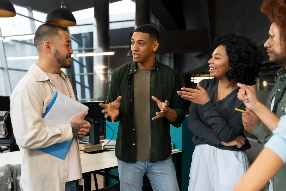 Grupo diverso de cinco pessoas em pé em um escritório moderno, engajados em uma conversa animada. Todos estão sorrindo, com expressões de entusiasmo e atenção. Um homem segura pastas com documentos enquanto os outros gesticulam durante a troca de ideias, transmitindo um clima colaborativo e positivo no ambiente de trabalho.