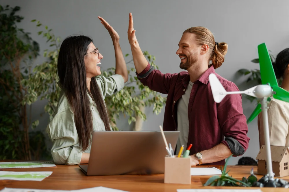 Dois colegas de trabalho, um homem e uma mulher, comemoram com um high-five em um ambiente de escritório sustentável. Eles sorriem enquanto trabalham em um projeto, rodeados por plantas e elementos ecológicos, como um pequeno modelo de turbina eólica sobre a mesa.