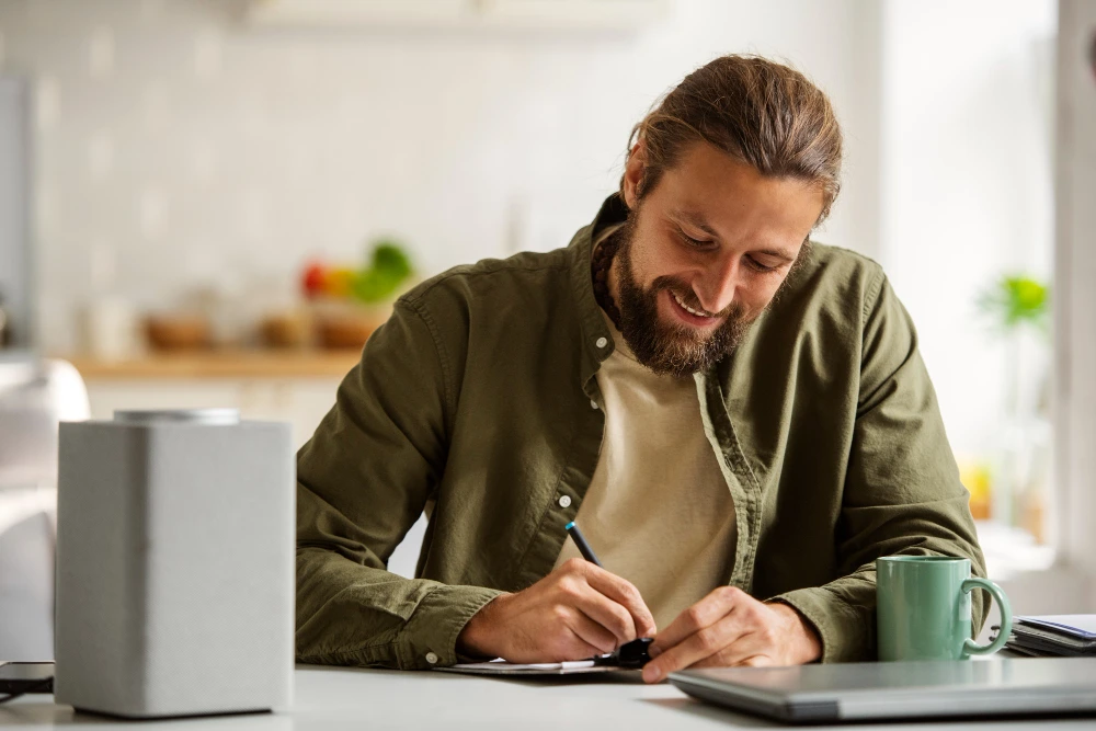 Homem jovem com barba e cabelo preso sorrindo enquanto escreve em um caderno sobre uma mesa branca, em um ambiente doméstico iluminado. Ao lado, há uma xícara verde, um laptop fechado e uma caixa de som. Ao fundo, a cozinha está desfocada, com utensílios e plantas decorativas. A cena transmite foco e bem-estar durante o trabalho remoto ou estudo.
