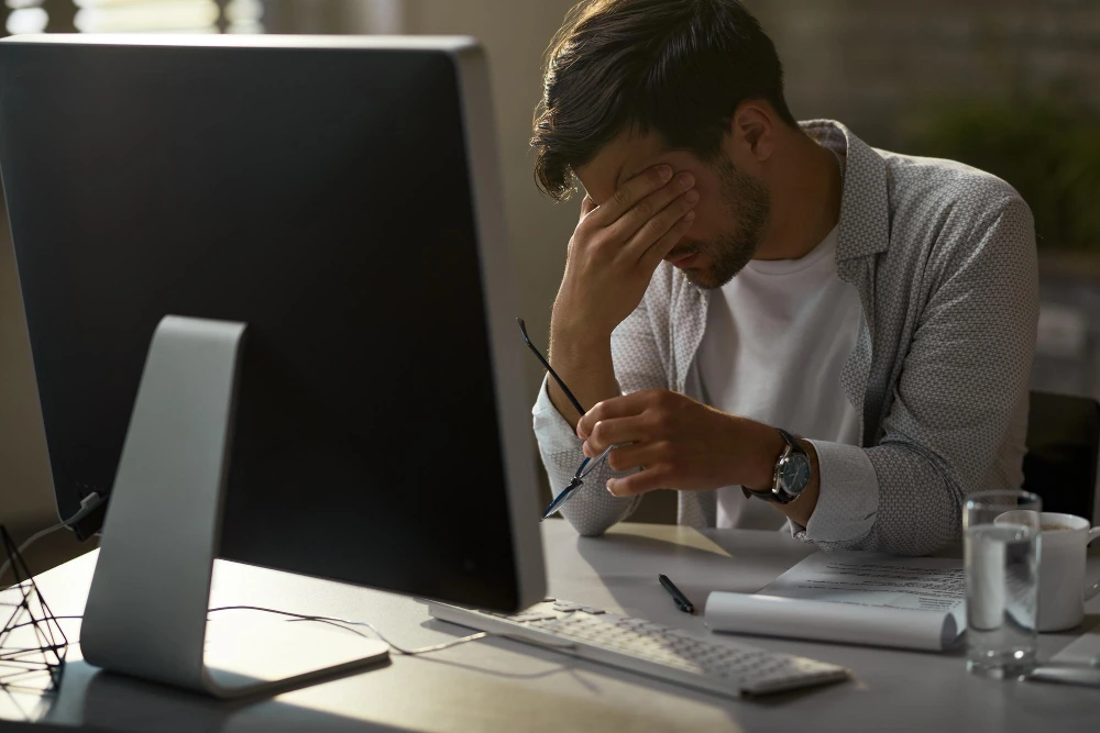 Homem sentado à frente de um computador grande, com expressão de cansaço ou estresse. Ele apoia o rosto em uma das mãos enquanto segura os óculos na outra. Sobre a mesa há um teclado, copo de água, caneca, papéis e uma caneta. A iluminação suave e o ambiente de escritório sugerem uma rotina intensa de trabalho ou esgotamento mental.
