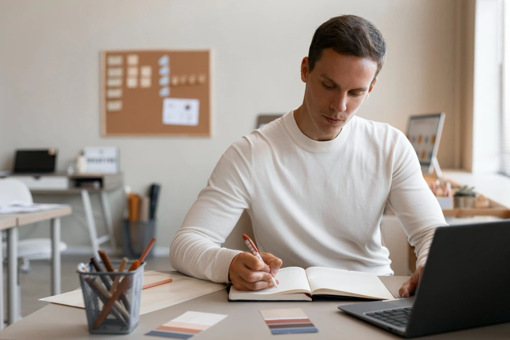 Homem jovem de cabelos curtos e camiseta branca está sentado à mesa em um ambiente de escritório, escrevendo em um caderno com um lápis. À sua frente, há um notebook aberto, amostras de cores e um porta-lápis. Ao fundo, é possível ver uma lousa de cortiça na parede e outros itens de papelaria em um ambiente bem iluminado.