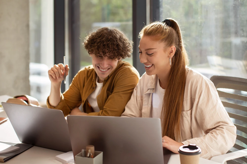 Dois jovens sorridentes trabalham lado a lado em um ambiente iluminado por luz natural. Ambos usam laptops e parecem compartilhar uma conversa descontraída. O homem, de cabelo cacheado e castanho, segura uma caneta enquanto observa a tela. A mulher, de cabelo ruivo e preso em rabo de cavalo, olha para o próprio notebook. Sobre a mesa há café e materiais de escritório, sugerindo um momento colaborativo e produtivo.