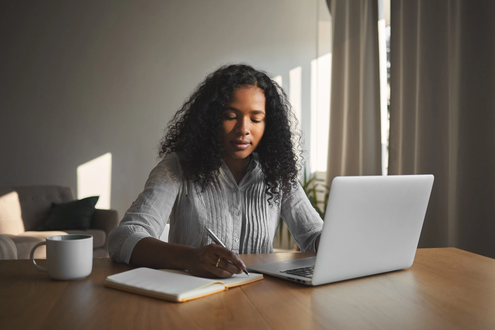 Mulher jovem de cabelos cacheados trabalhando em frente a um laptop em casa, fazendo anotações em um caderno, com uma xícara ao lado sobre a mesa de madeira.