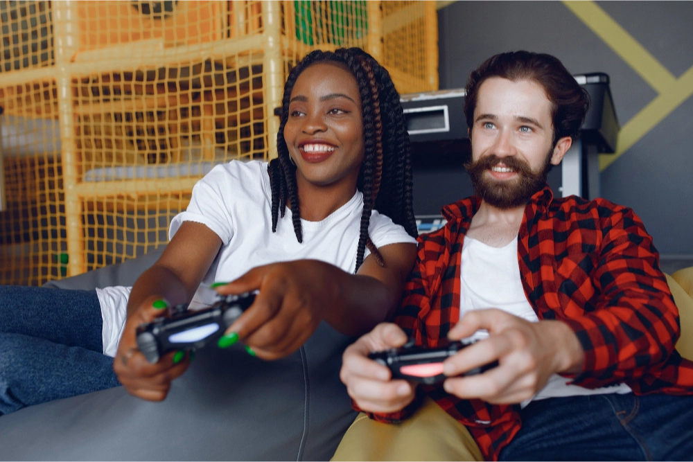 Duas pessoas sorridentes, uma mulher e um homem, jogando videogame juntas em um ambiente descontraído, segurando controles de console.
