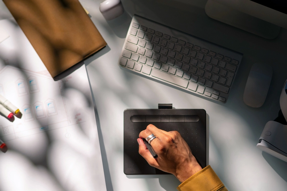 Pessoa utilizando uma mesa digitalizadora em uma estação de trabalho, com teclado, mouse e materiais de escritório ao redor.