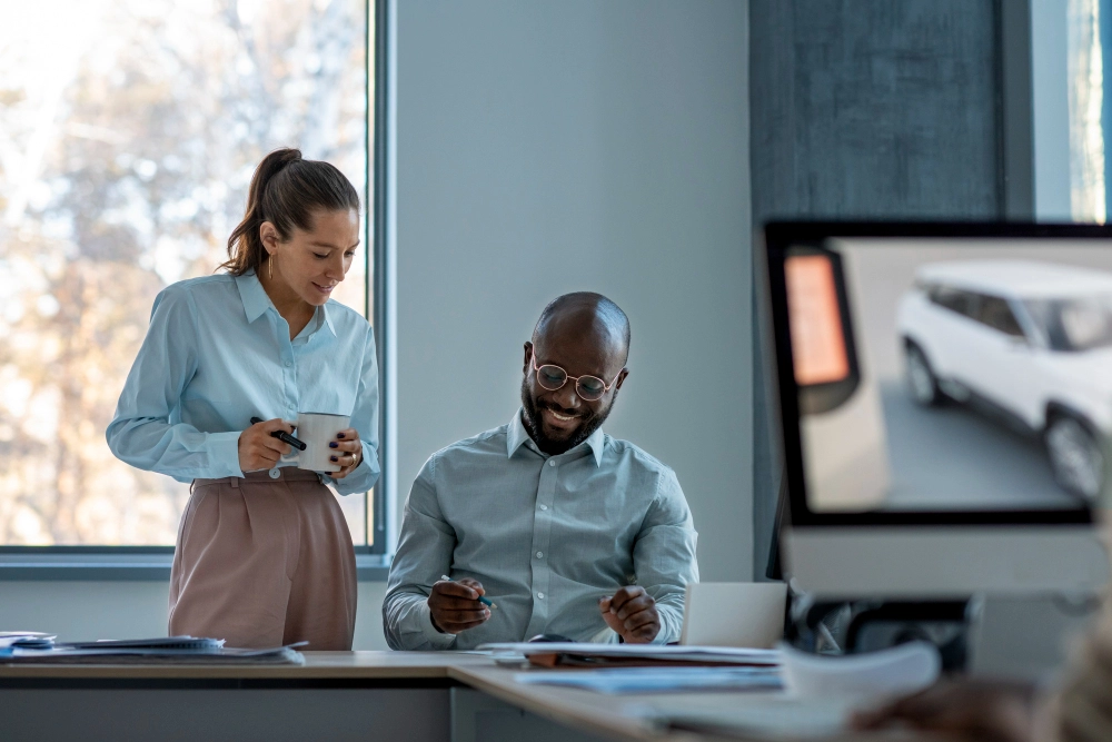 Dois colegas de trabalho em um escritório iluminado pela luz natural. Um homem sentado, sorrindo enquanto analisa documentos, e uma mulher em pé ao seu lado, segurando uma caneca e uma caneta, observando o material. Ao fundo, uma janela ampla e um monitor de computador exibindo a imagem de um carro.
